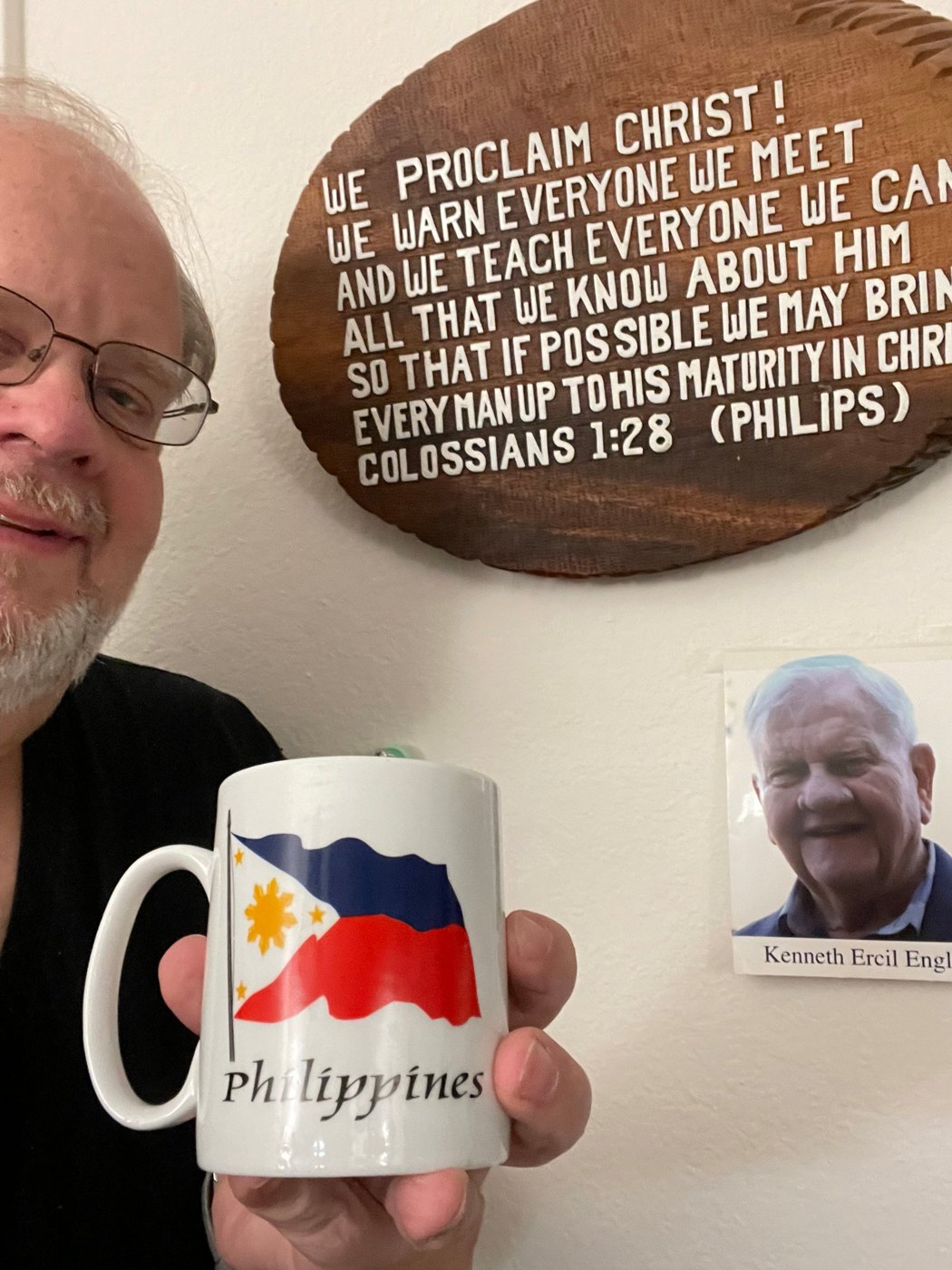 Kevin Engle holding a Philippines mug beneath Dad's Colossians 1:28 plaque and his memorial photo