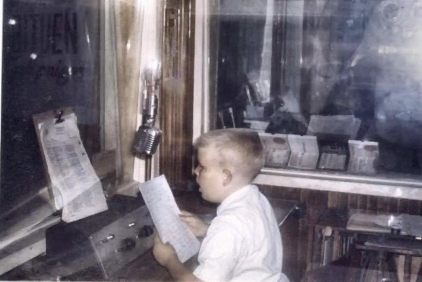 Kevin Engle as a boy, reading a script at a station microphone in a radio booth, with the control room behind the glass