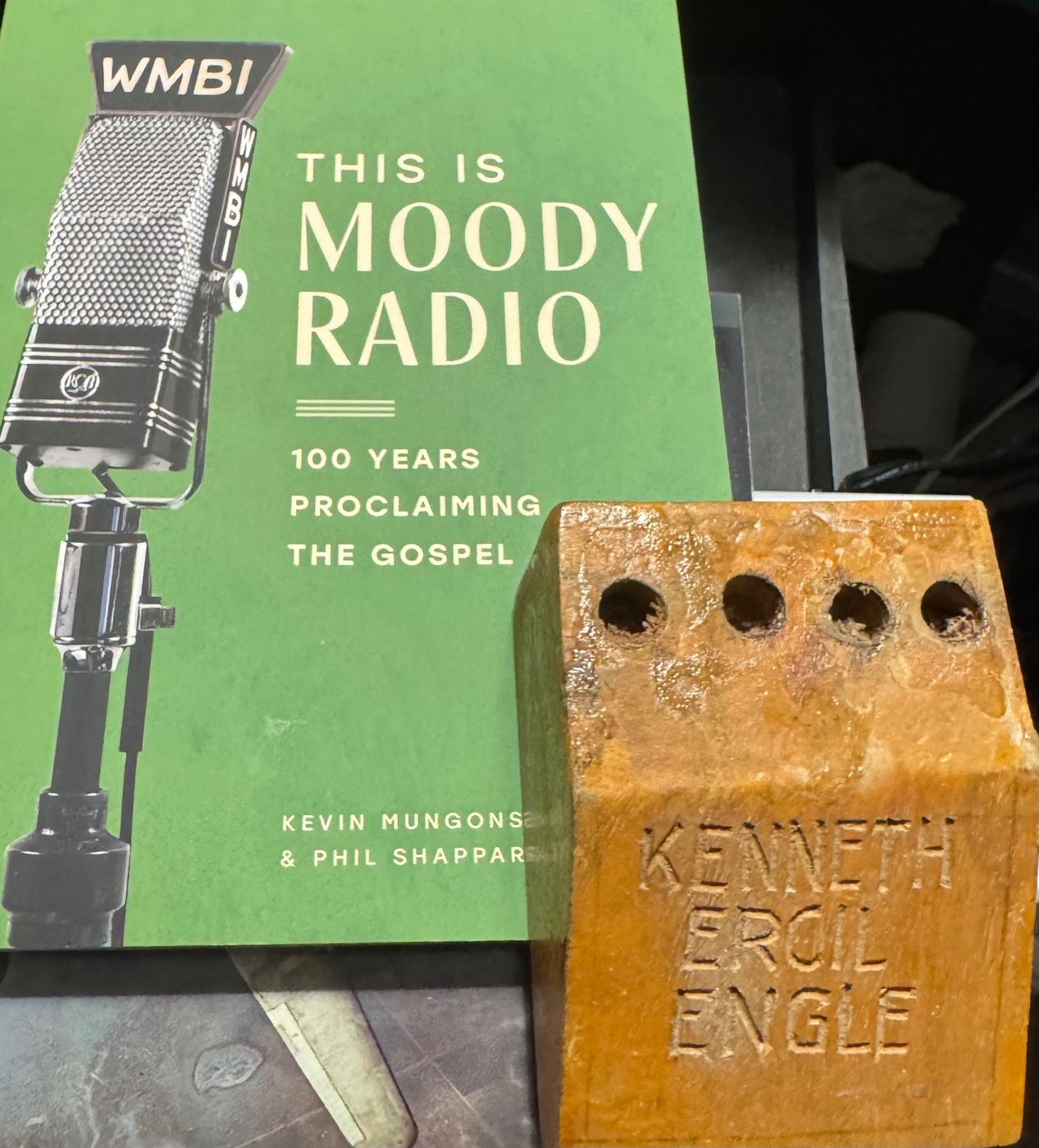 A wooden pencil holder carved with the name KENNETH ERCIL ENGLE, standing in front of the book This Is Moody Radio: 100 Years Proclaiming the Gospel