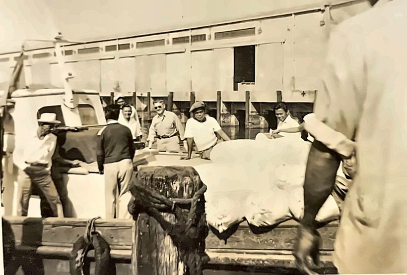 Men on a Manila pier beside a launch loaded with canvas-covered cargo, a pier shed behind them, March 1951