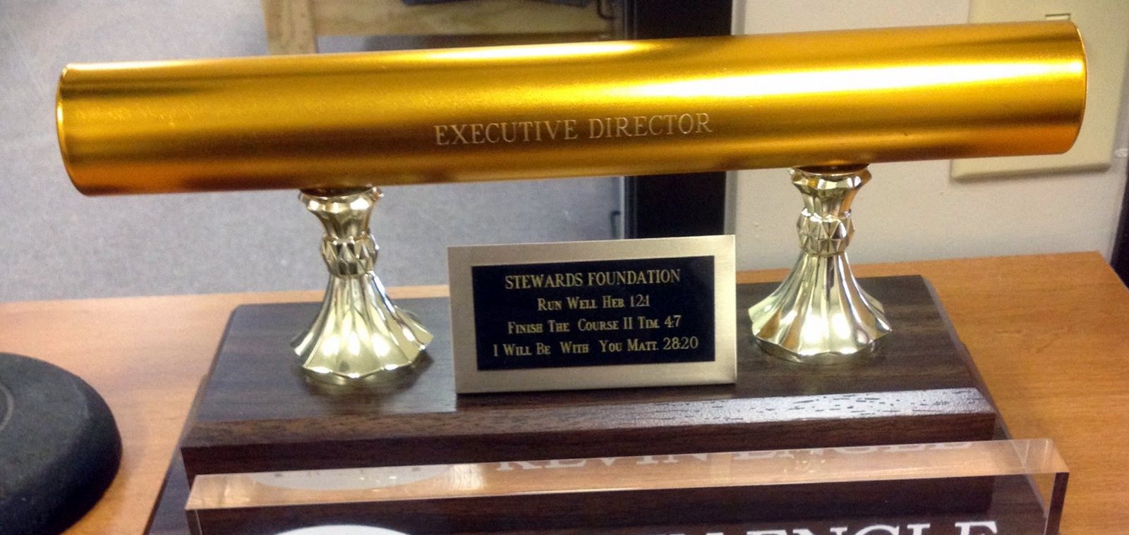 The gold Executive Director baton that trustee Jerry Keller presented to Kevin when he went off the board in 2006, in grateful thanks for Kevin's years of work with Stewards Foundation. The plate reads: Run Well, Heb. 12:1 · Finish the Course, II Tim. 4:7 · I Will Be With You, Matt. 28:20.